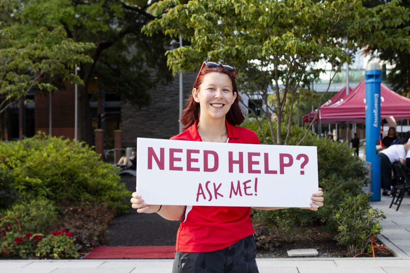Woman in cherry holds a sign saying "Needs help? Ask me!"